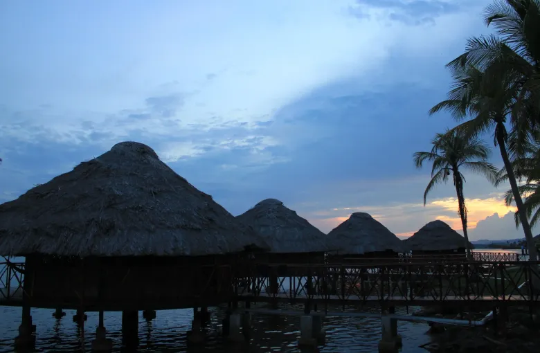 Thatched cabins standing on stilts over calm water in Guna Yala at dusk, with palms behind.