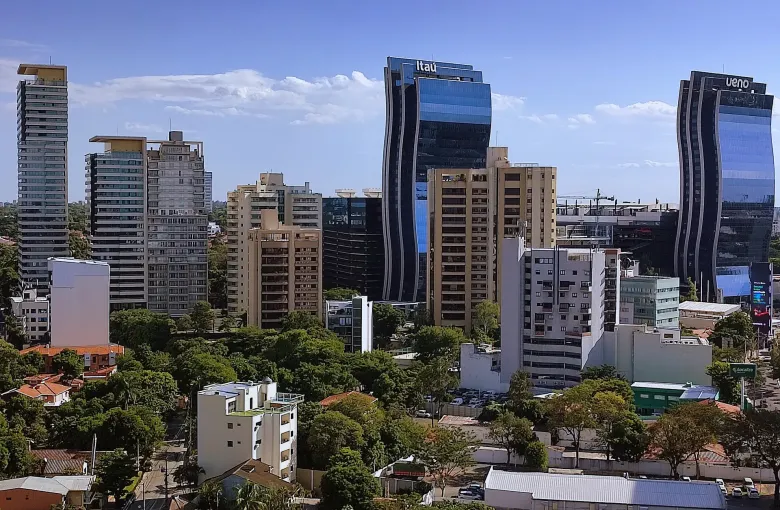 Glass bank towers rise above tiled rooftops and dense green trees in the business district of Asuncion.