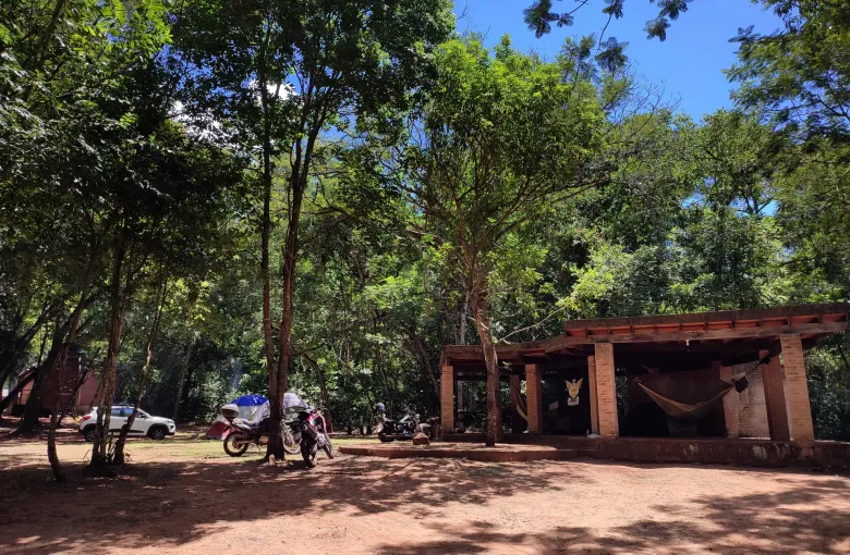 Hammocks hang under a brick shelter beside motorcycles and red dust at a campsite in Cerro Cora National Park.