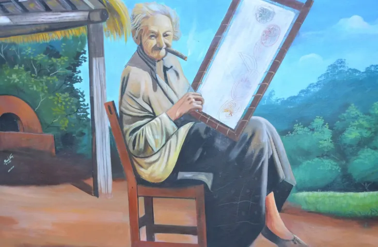 A painted wall mural shows a woman working a nanduti lace web stretched on its round wooden frame.