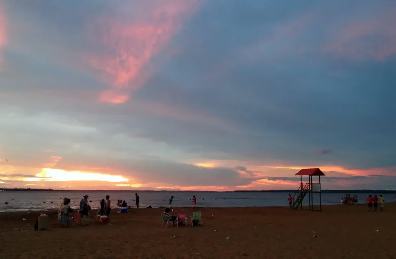 Families gather on the sand at Encarnacion as a pink and orange dusk settles over the Parana river.