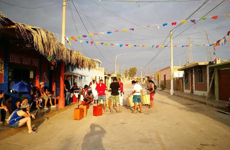 Wooden cajon drums set out in a street at El Carmen in Chincha under lines of paper bunting, with people gathering to play.