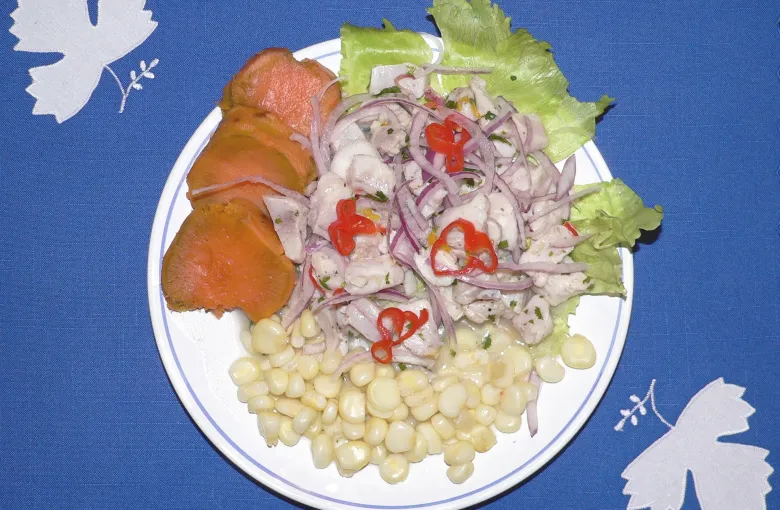 A plate of Peruvian ceviche with sliced red onion and chilli, sweet potato, choclo corn and lettuce.