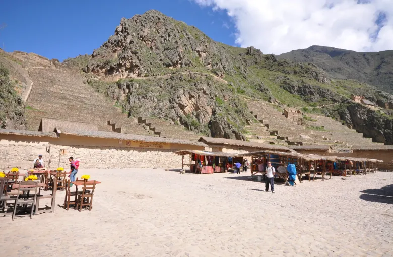 Rows of covered craft stalls on the open square at Ollantaytambo with the Inca terraces climbing the hillside behind.