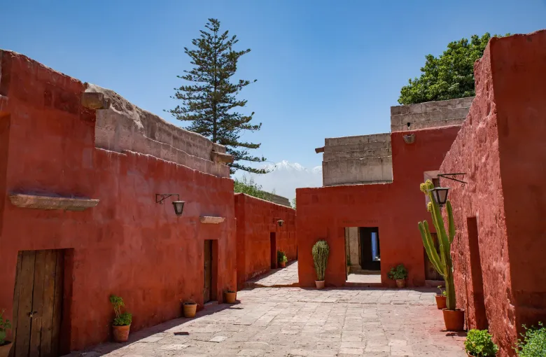 A lane between deep red walls inside the Santa Catalina Monastery in Arequipa, with doorways and a tree above the roofline.