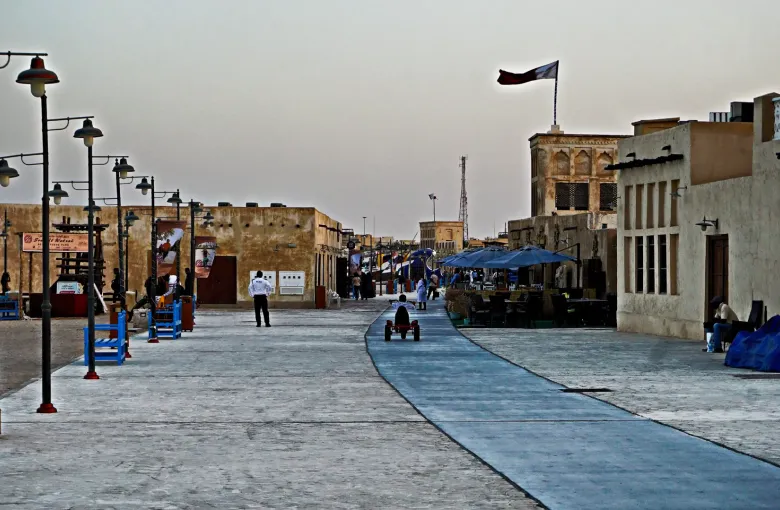 A paved waterfront promenade at Souq Al Wakrah lined with coral stone buildings and a Qatari flag
