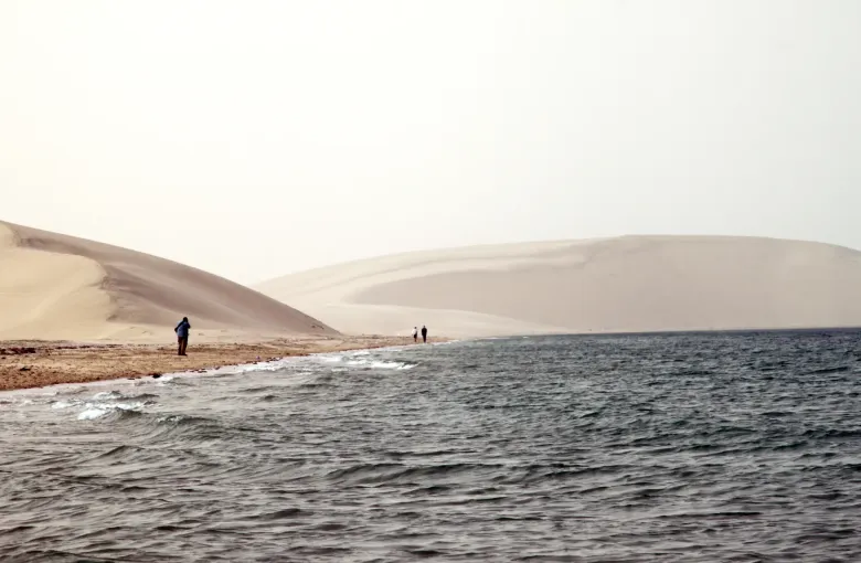 Pale dunes running down to the shallow water of the inland sea at Khor Al Adaid, with two figures at the waterline