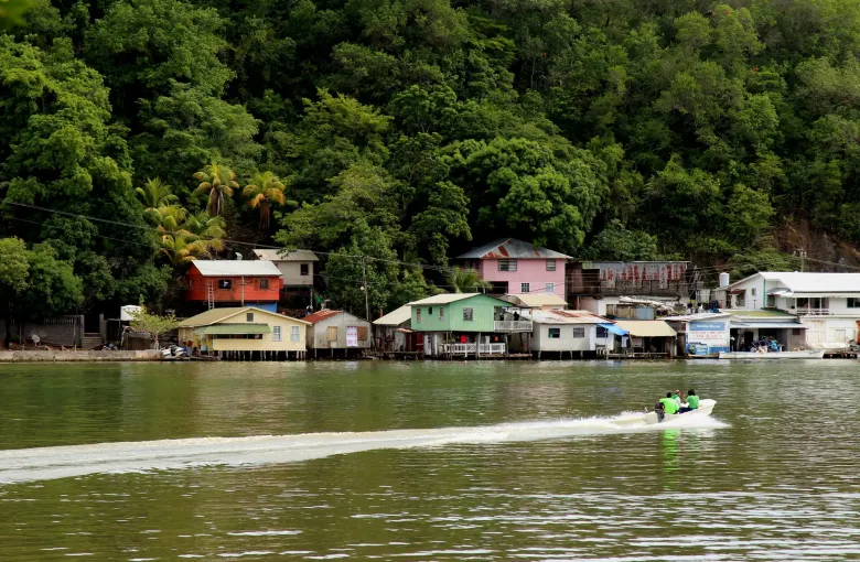 A small open boat under way past timber houses built on stilts over the water at Oak Ridge.
