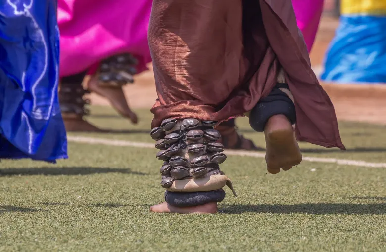 Close view of Intore dancers mid-step, seed-pod amayugi bells bound at their ankles over brightly colored wraps, on a pitch in Rwanda.