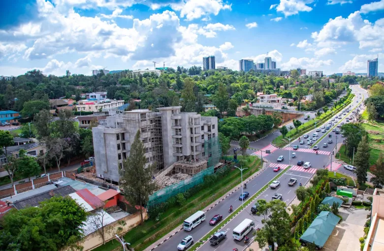 A divided boulevard in Kigali seen from above, with minibuses, motorcycle taxis and cars, a building under construction, and the downtown towers on the ridge behind.