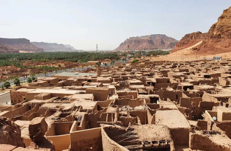 The mud-brick houses of AlUla old town below a sandstone escarpment, with the palm oasis stretching away behind them.