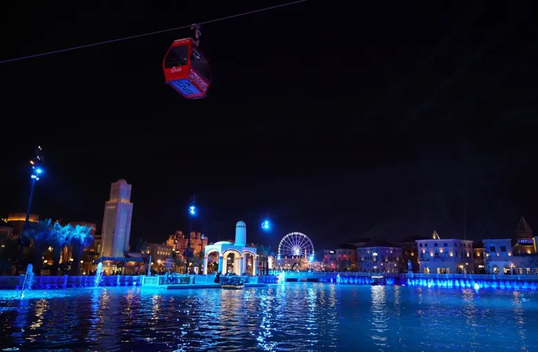 The lit waterfront promenade of Boulevard City in Riyadh, Saudi Arabia, with a cable car crossing above the paved walkways.