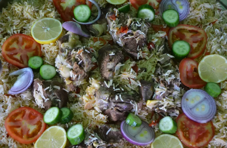 A shared platter of kabsa, spiced rice with meat, dressed with sliced tomato, cucumber, onion and lemon.