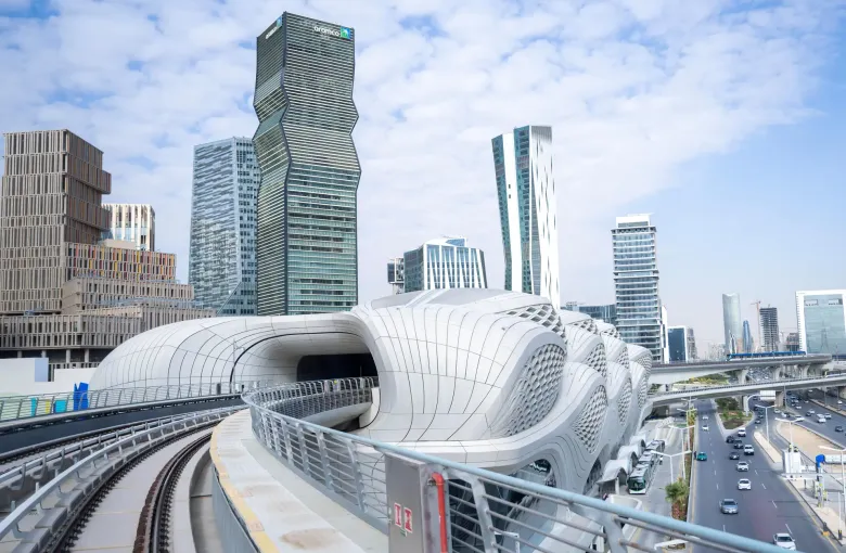 A Riyadh Metro station in Saudi Arabia, its curved white roof open to the track with the towers of the financial district behind.