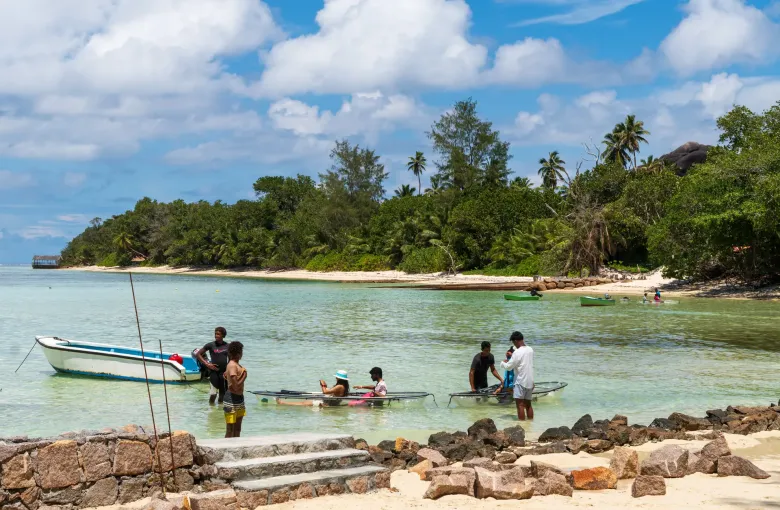 Small craft drawn up on a sheltered shore at La Digue, people wading in the shallows and a wooded headland beyond.