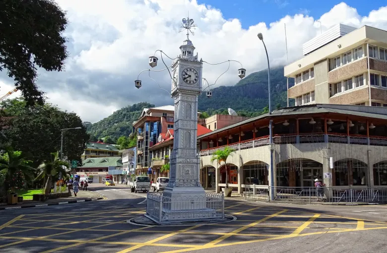 The silver clock tower at the central junction of Victoria on Mahé, colonial-era buildings and wooded hills behind.