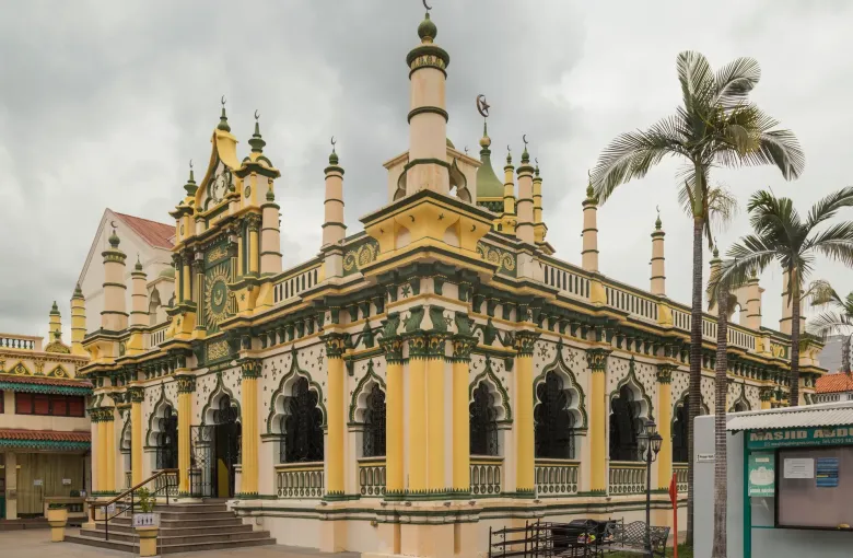The ornate yellow and white facade of the Abdul Gafoor Mosque in Little India, Singapore, with palms against a cloudy sky.