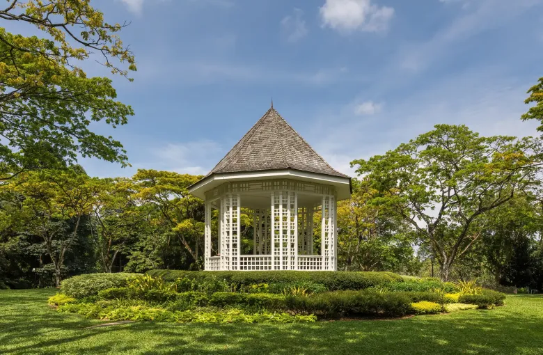 The white octagonal bandstand on its mown rise among mature trees at the Singapore Botanic Gardens on a sunny afternoon.