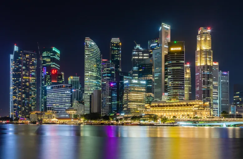 The lit towers of Singapore's central business district reflected in the water of Marina Bay at night.