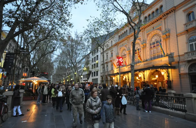 A dense evening crowd on the tree lined promenade of La Rambla in Barcelona, Spain, the lit Liceu theater beyond.