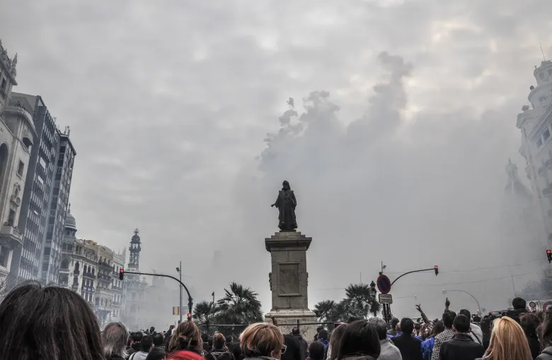 Firework smoke drifting over a statue and a packed crowd in the city square during a mascleta at Las Fallas in Valencia.