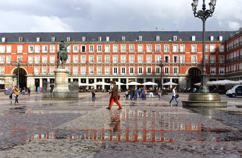 Wet cobbles reflecting the red arcaded houses of the Plaza Mayor in Madrid, Spain, after rain, an umbrella at the edge.