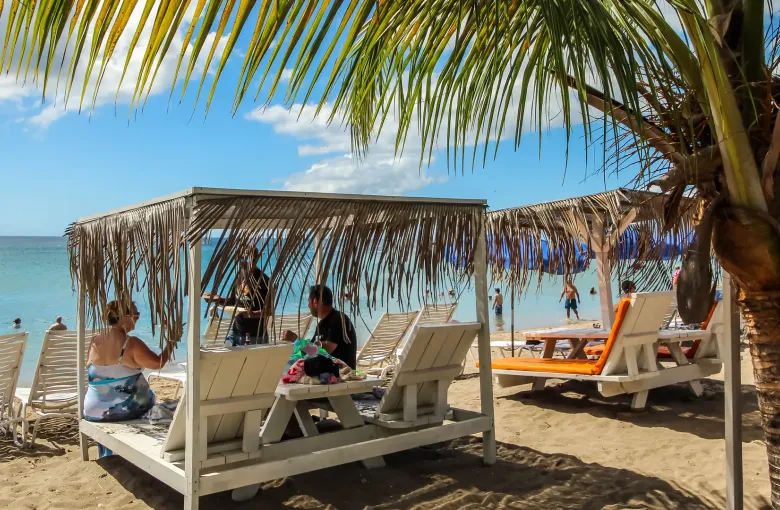 Thatched shade structures and wooden loungers on the sand at Pinney's Beach on Nevis, with swimmers in calm water beyond.