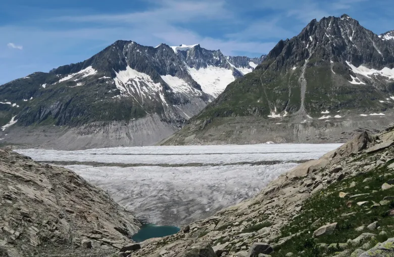 The dirty snout of the Aletsch Glacier between bare rock walls, with a small green meltwater lake collecting below it.