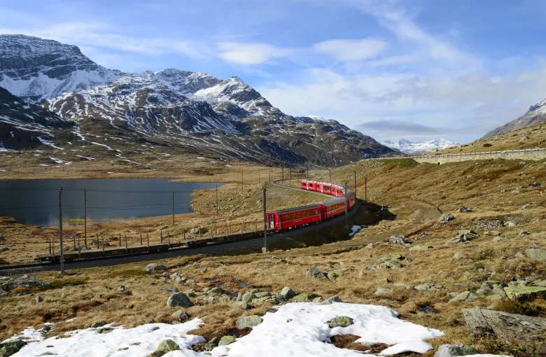A red regional train running along the shore of Lago Bianco on the Bernina line, snow still lying on the high pasture.
