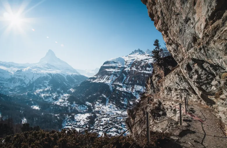 The Matterhorn and the snow-covered village of Zermatt seen out from under a bare rock overhang on the slope above.