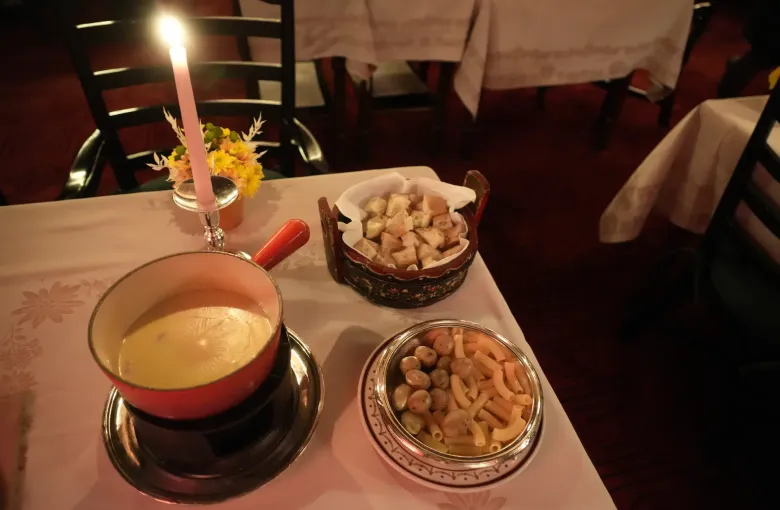A caquelon of Swiss cheese fondue on its burner with bowls of cut bread and boiled potatoes beside it on a white cloth.
