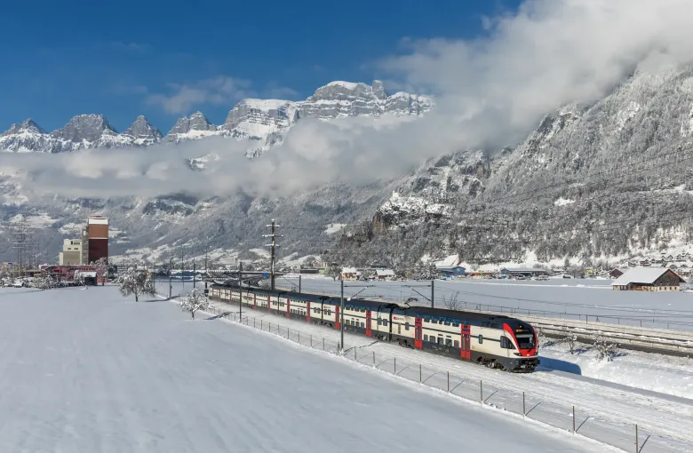 A modern Swiss double-deck train running through snow-covered fields with cloud lying on the peaks behind.