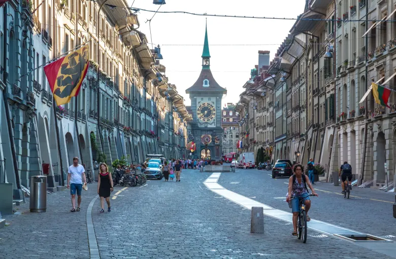 The Zytglogge clock tower at the end of a cobbled street in Bern's old city, with arcades and cantonal flags either side.
