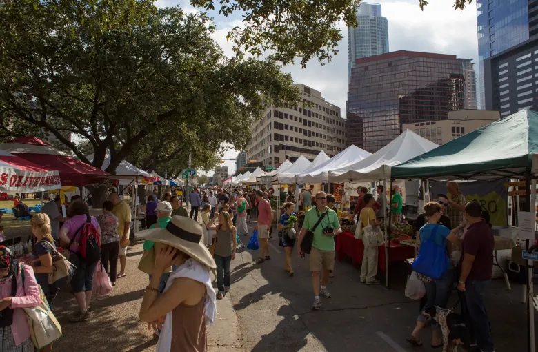 Shoppers browse white canopy stalls under live oaks at an outdoor farmers market, with downtown Austin towers behind.