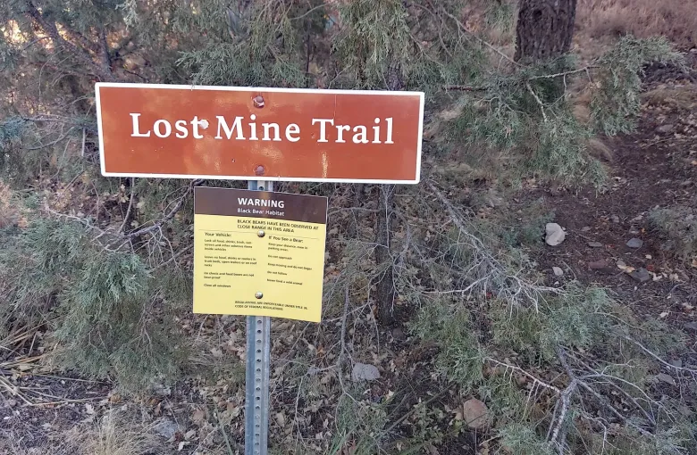 A brown Lost Mine Trail sign above a yellow warning notice about black bear habitat, set in dry juniper scrub.