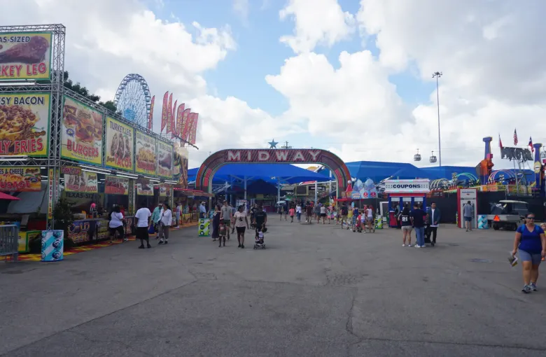 Fairgoers walk toward a red Midway arch past food stands, with a tall Ferris wheel and blue canopies under a cloudy sky.