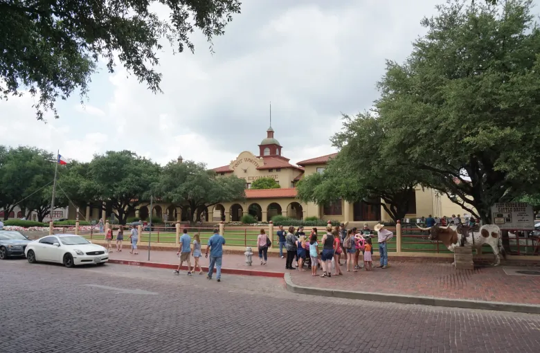 Visitors gather on a brick street beside a saddled longhorn, in front of a mission style exchange building with a cupola.