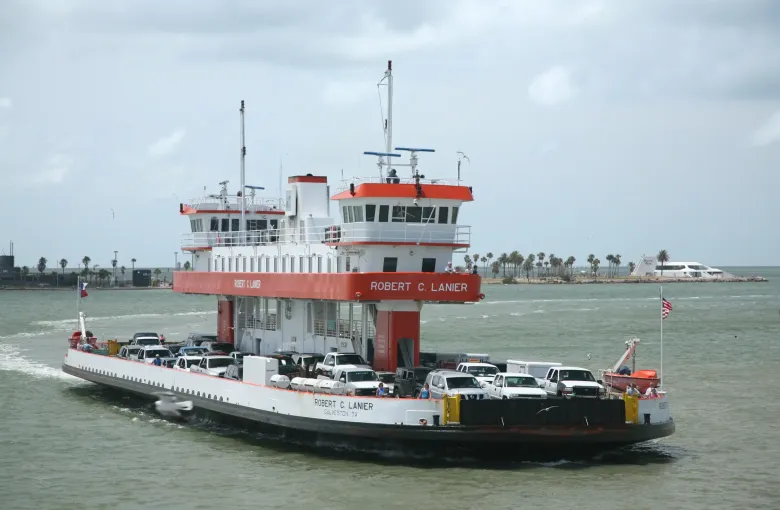 A white and red car ferry loaded with pickups and cars crosses gray green water, with palm trees on the shore behind.