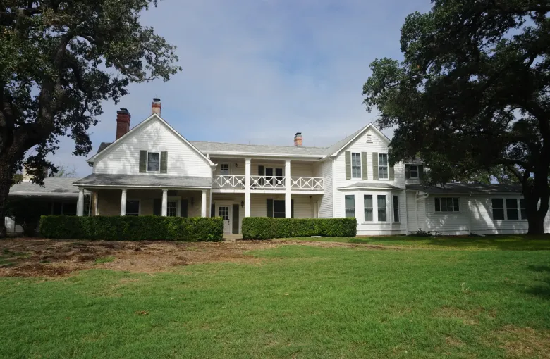 A long white two-story ranch house with a columned porch and green shutters, shaded by old live oaks over a lawn.