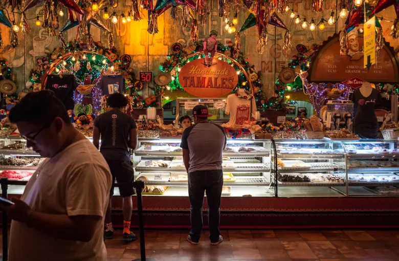 Customers wait at a long glass bakery case under a Handmade Tamales sign, strings of lights and bright tinsel decorations.
