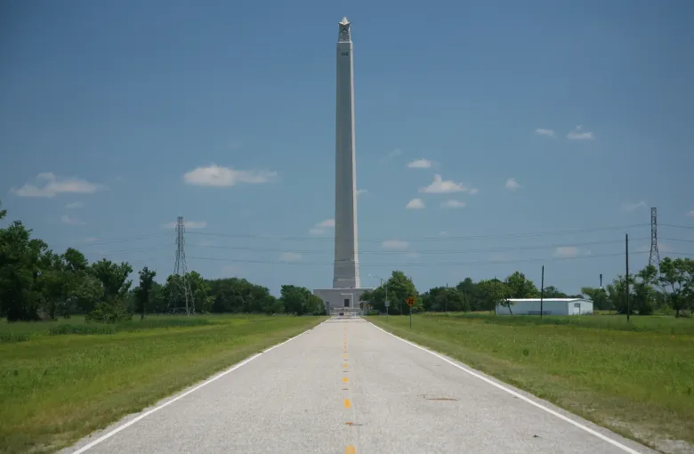 A tall stone column topped with a star rises at the end of a straight road through flat green grassland.