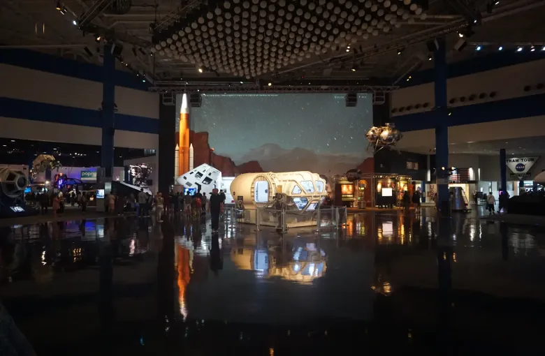 Visitors cross a wide, level exhibit hall with a polished floor, spacecraft models and a rocket mural lit against the dark.