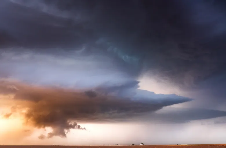 A dark, layered supercell thunderstorm cloud spreading over a plowed brown field, with a band of orange light beneath it.