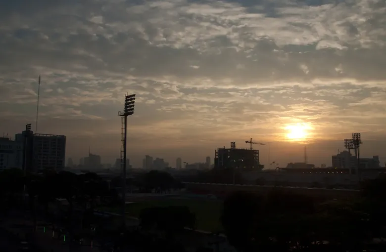 The sun low behind heavy cloud over the rooftops and towers of Bangkok, seen from an elevated station.