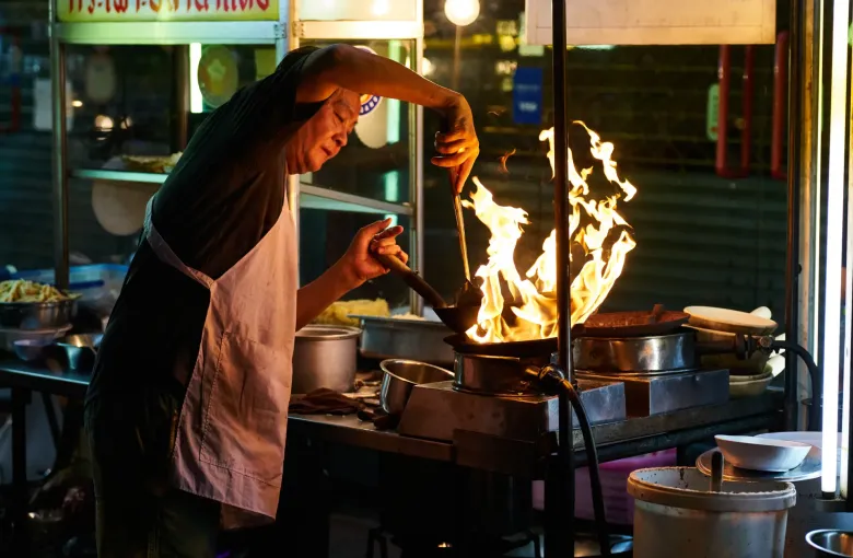A cook leaning over a wok that has caught flame at a lit street food stall in Bangkok after dark.