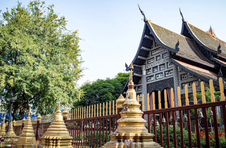 A dark timber Lanna temple in Chiang Mai behind a row of small gilded chedi on a level paved forecourt.