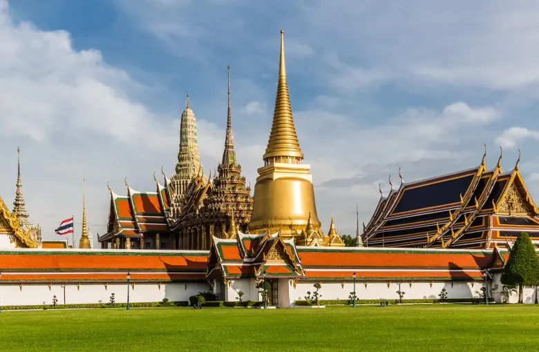 Gilded spires and tiled roofs of the Grand Palace in Bangkok rising behind a lawn under a bright blue sky.