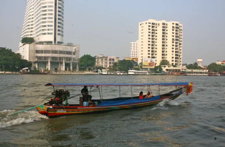 A longtail boat with a canopy carrying passengers along the river in Bangkok, buildings on the far bank.