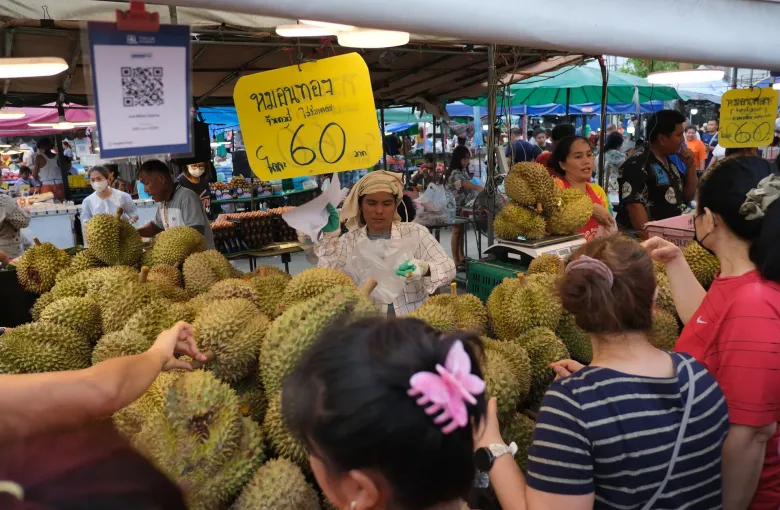 Shoppers around a market stall heaped with durians, a yellow sign giving the price in baht above them.