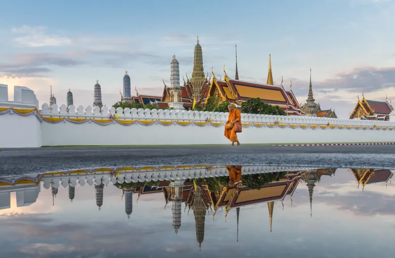 A monk in orange robes walking beside the white wall of Wat Phra Kaew, the temple spires reflected in water.